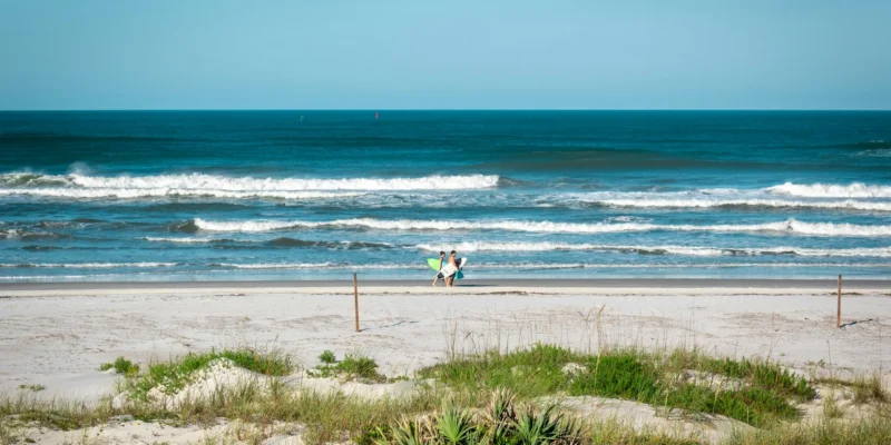 Two people carrying surfboards on New Smyrna Beach, with breaking waves behind them